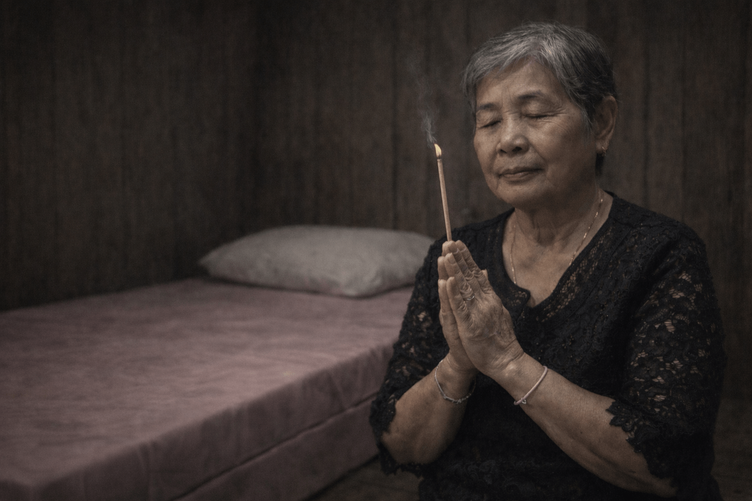 หญิงสูงวัยชาวเอเชียนั่งพนมมือถือธูป 1 ดอก หลับตาอย่างสงบ อยู่ข้างที่นอนสีชมพูในห้องไม้แสงสลัว บนเตียงมีหมอนวางเรียบร้อย ไม่มีผ้าห่มของพระ บรรยากาศเงียบสงบและอบอุ่น