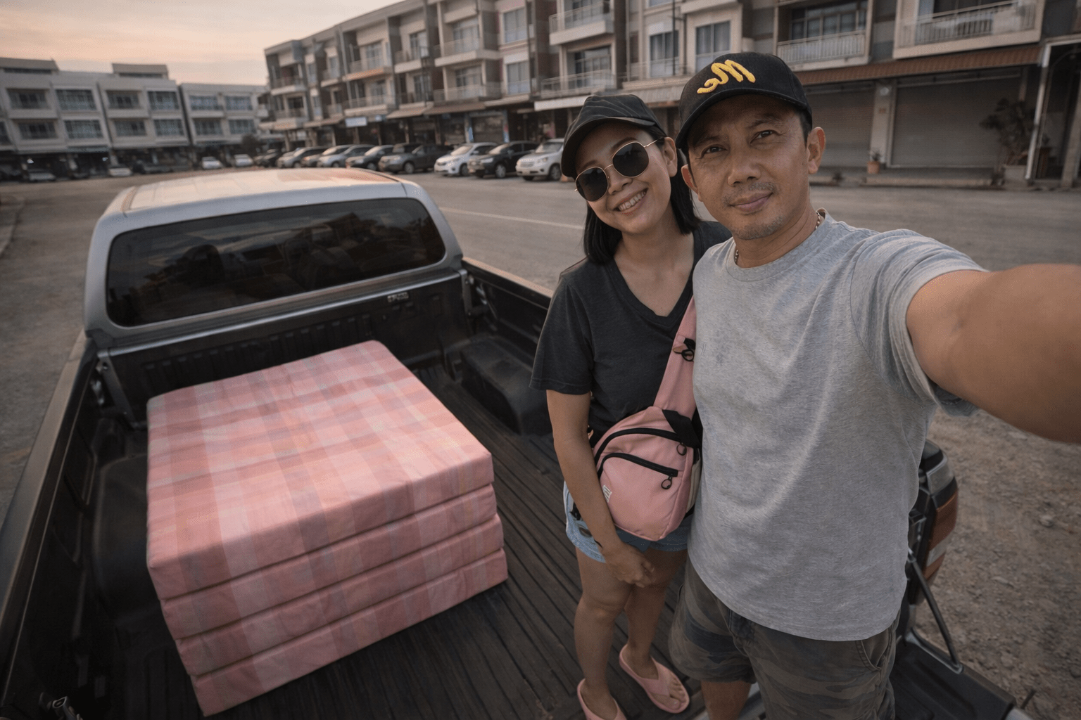 ภาพถ่ายแนวนอนมุมสูงของผู้ชายและผู้หญิงยืนอยู่ท้ายรถกระบะในช่วงเย็น ทั้งคู่ยิ้มเล็กน้อยคล้ายกำลังบอกลา ภายในกระบะมีที่นอนสีชมพูพับสี่ทบวางเรียบร้อย ฉากหลังเป็นอาคารพาณิชย์และถนนในแสงอาทิตย์ยามเย็นโทนอุ่น